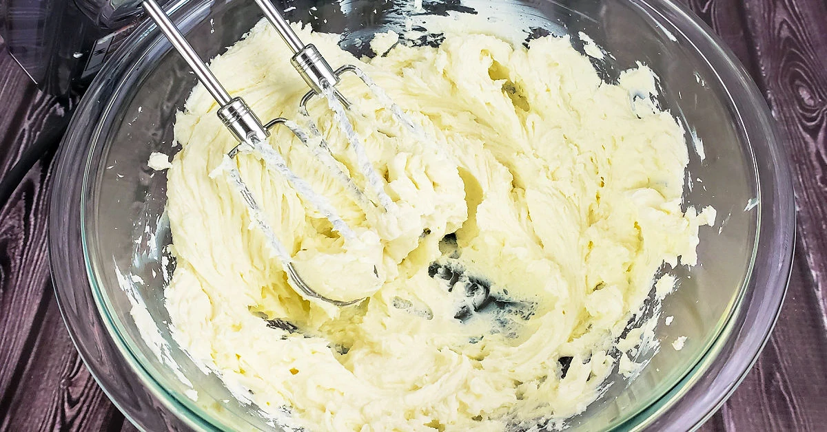 Cream cheese being beaten with an electric hand mixer in a glass mixing bowl.
