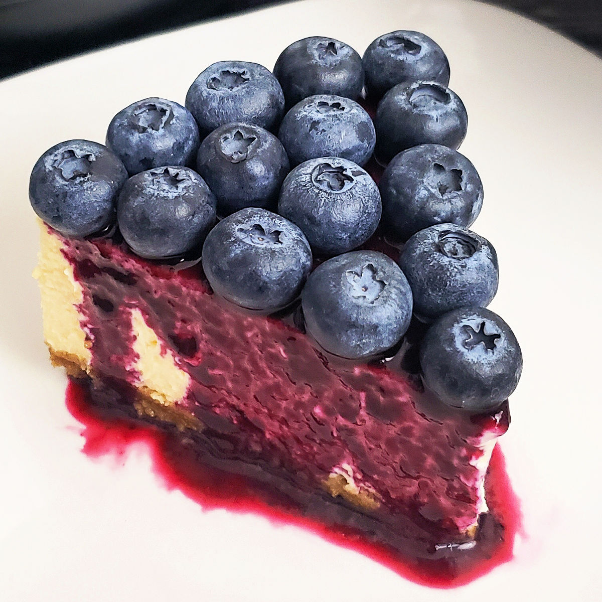 Slice of New York Style cheesecake topped with blueberry syrup and fresh blueberries.