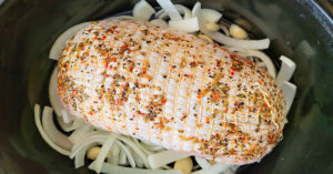 Seasoned boneless turkey roast with netting set on top of onions and garlic in a slow cooker.
