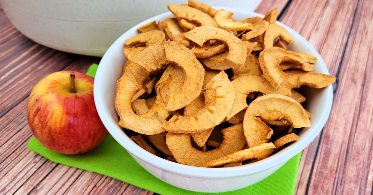 Bowl of dehydrated apple chips on green napkin next to a fresh apple and dehydrator.