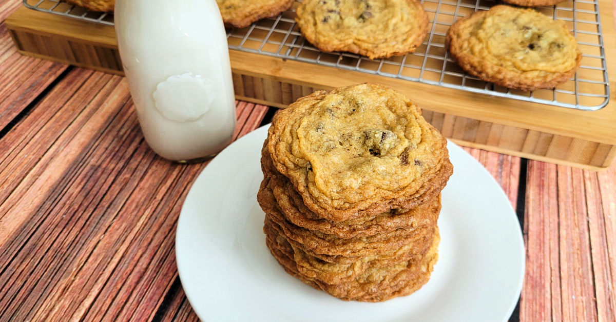Stack of gluten free chocolate chip cookies on a white dessert plate with a glass of milk in front of a rack of more cookies cooling.
