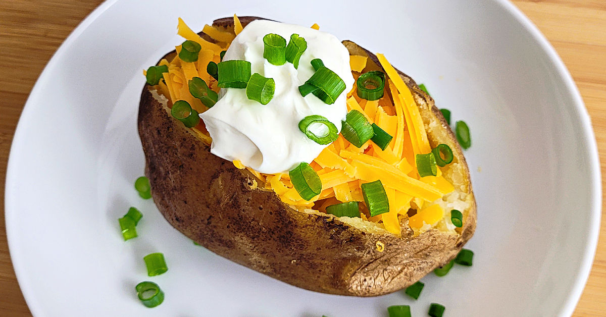 Slow cooker baked potato on a white plate, topped with cheese, sour cream and green onions.