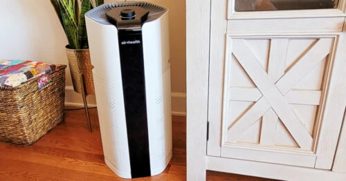Air Health Skye Air Purifier next to a white TV stand, tall green plant and basket of quilts on hardwood floors.