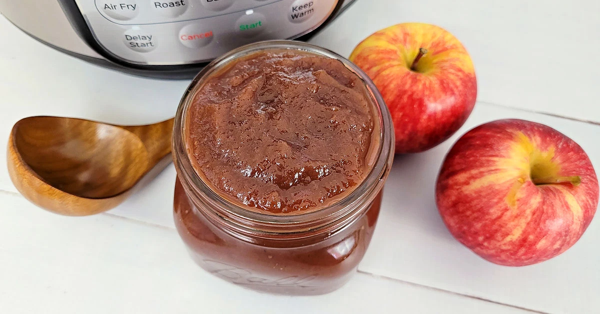Pint jar of apple butter surrounded by an Instant Pot, 2 apples and wooden ladle.